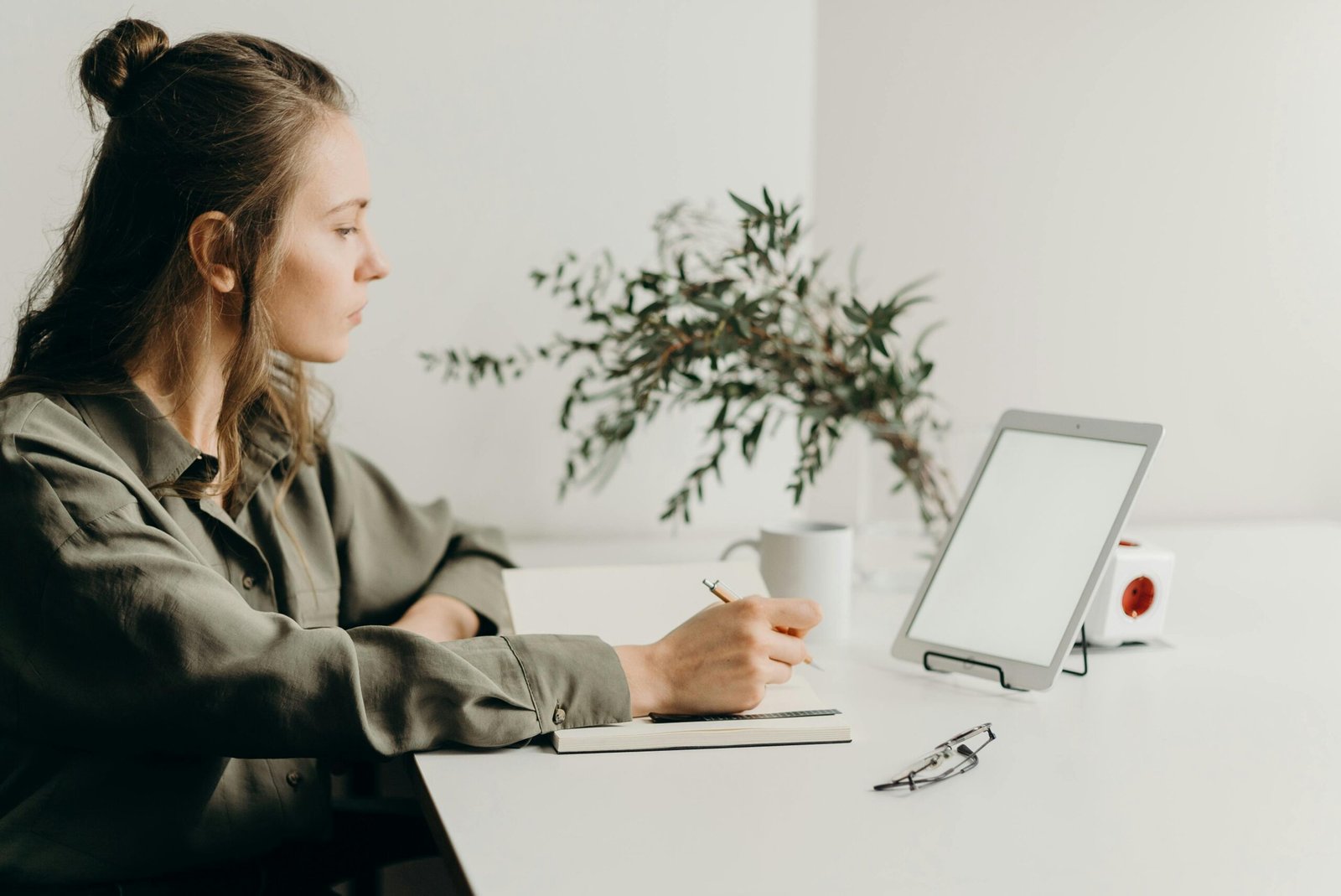 Freelancer writing notes in a minimalist home office with a white tablet, plant, and reading glasses.